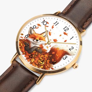 May include: A gold-tone wristwatch with a brown leather band. The watch face features a smiling fox nestled in autumn leaves. The watch hands point to 10:10. The watch has a white background with black numerals.