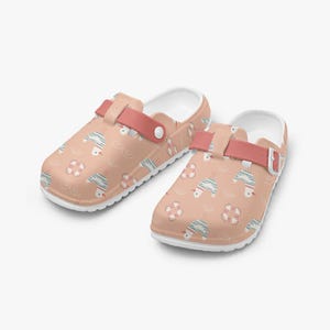 May include: Peach-coloured clogs with a white interior and a coral strap. The clogs feature a repeating pattern of seagulls, waves, and life preservers. The clogs have a white sole.