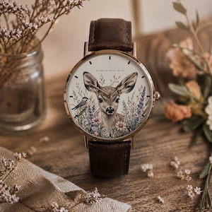 May include: A gold-toned wristwatch with a brown leather band. The watch face features a deer illustration surrounded by flowers and a small bird. The text "BE KIND. BE GENTLE. BE YOU." is printed on the watch face.