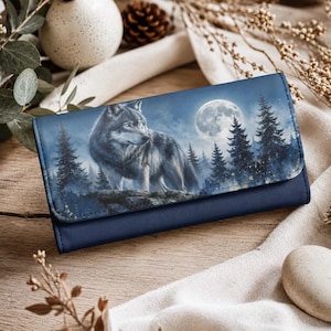 May include: A rectangular navy blue wallet featuring a printed graphic on the flap. The image depicts a grey wolf standing on a rocky outcrop within a dark forest of pine trees under a large, bright full moon against a deep blue night sky.