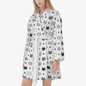 May include: White robe with a repeating pattern of black and gray cat faces, paw prints, and stars. The robe has a matching white belt and a shawl collar. The robe is knee-length and has long sleeves.