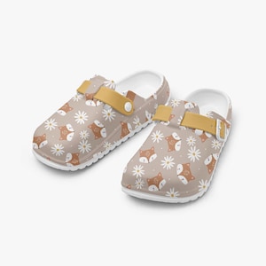 May include: A pair of clogs with a tan background, white daisies, and orange foxes. The clogs have white soles, tan straps, and a buckle closure. The clogs are designed for comfort and style.