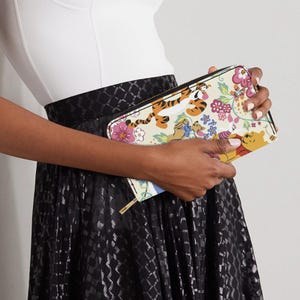 May include: A cream-colored zip-around wallet featuring a colorful floral and cartoon character design. The design includes images of Tigger, Winnie the Pooh, Piglet, and Eeyore. The wallet has a gold-colored zipper.