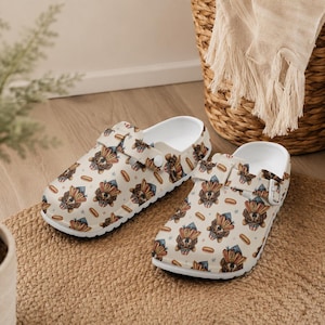 May include: A pair of white clogs with a repeating pattern of a dog wearing a patriotic hat, hot dogs, and stars. The clogs have a white interior and a strap that can be adjusted. The shoes are on a woven mat.