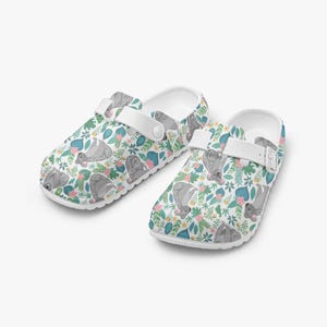 Pode incluir: Tamancos brancos com um padrão colorido de bichos-preguiça cinzentos, flores cor-de-rosa e folhas verdes. Os tamancos têm tiras brancas e sola branca. Estes sapatos são perfeitos para uso casual.