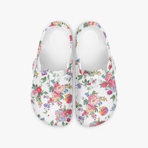 May include: A pair of white clogs adorned with a colourful floral print. The pattern showcases pink, red, yellow, and blue flowers with green foliage. These clogs have a classic slip-on style and a pivoting heel strap.