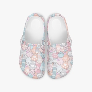 May include: A pair of white clogs with a colorful pattern of cat paws in pastel shades of pink, blue, gray, and peach. The clogs have a rounded toe and a strap that can be worn around the heel.