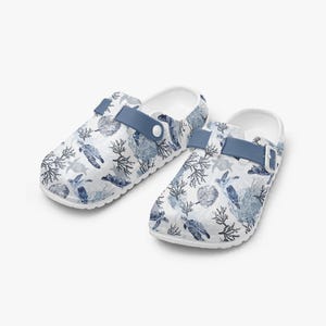 Könnte beinhalten: Weiße Clogs mit einem blau-weißen Meeresschildkröten- und Korallenmuster. Die Clogs haben einen blauen Riemen und eine weiße Sohle. Diese Schlupfschuhe sind auf Komfort und Stil ausgelegt.