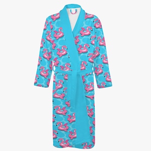 May include: A turquoise bathrobe with a pattern of pink flamingo pool floats. The robe has a shawl collar, long sleeves, and a matching belt. The background is a lighter turquoise with white polka dots.