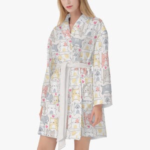 May include: White robe with a repeating pattern of cartoon cats in various colors, including pink, yellow, and gray, with small pink hearts. The robe has a white belt and a shawl collar.