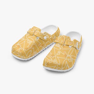 May include: A pair of yellow clogs with a white outline cat pattern. The clogs have white soles and adjustable straps. The shoes are designed for comfort and casual wear.