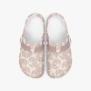 Peut inclure: Paire de sabots beiges avec un motif de coquillages beige foncé. L'intérieur est blanc et la bride et la boucle sont mauves. Ces sabots sont conçus pour un usage décontracté et confortable.