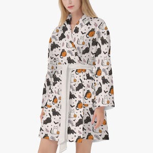 May include: White robe with a repeating pattern of black cats, orange pumpkins, fish bones, and bats. The robe has a white belt and is knee-length. The pattern is in black, orange, and pink on a white background.