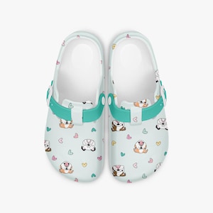 May include: A pair of light blue clogs with a repeating pattern of cartoon badgers and hearts in various colors. The clogs have a white interior, teal straps, and teal buckles.
