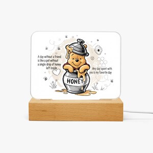 May include: A decorative night light featuring a cartoon bear character sitting in a honey pot. The light has a wooden base and a white panel with the text "A day without a friend is like a pot without a single drop of honey left inside."
