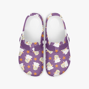May include: A pair of purple clogs with a white interior and a pivoting heel strap. The clogs feature a repeating pattern of cartoon ghosts and orange stars. The clogs are designed for comfort and casual wear.