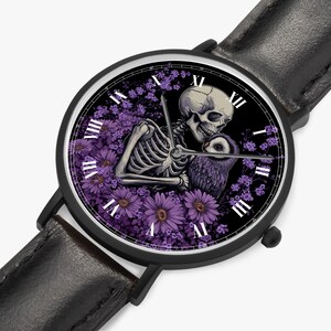 May include: Black wristwatch with a black leather band. The watch face features a skeleton embracing an owl amidst purple flowers against a black background. Roman numerals mark the hours.