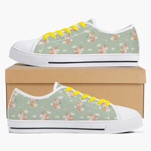 May include: Canvas trainers featuring a repeating pattern of orange foxes and white daisies on a pale green backdrop. The shoes have white soles, white toe caps, and yellow laces. The trainers are displayed on a brown cardboard box.