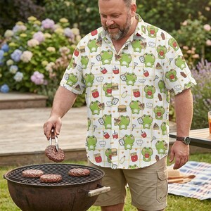May include: A short-sleeved button-down shirt with a white background and a repeating pattern of green frogs, burgers, fries, and drinks. The shirt is worn by a person grilling burgers on a charcoal grill.