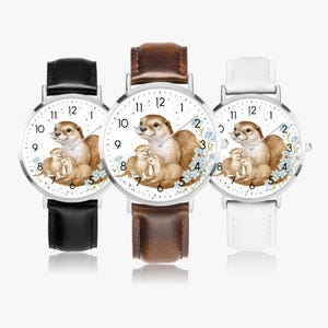 May include: Three wristwatches with silver cases and leather straps. The watch faces feature an illustration of otters and blue flowers. The straps are black, brown, and white. The watch face has black numbers.