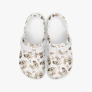 May include: White clogs featuring a repeating pattern of watercolour chickens in brown, grey, and yellow. The clogs have a white strap and a moulded footbed. These clogs are designed for comfort and casual wear.