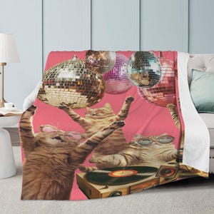 May include: A pink throw blanket featuring a whimsical design of cats wearing sunglasses and holding disco balls. One cat is operating a record player. The blanket is draped over a couch, creating a fun and playful aesthetic.