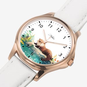 May include: A white leather wristwatch with a rose gold case and a white face. The face features a watercolour illustration of an otter in a river, with Roman numeral hour markers and a black minute hand.