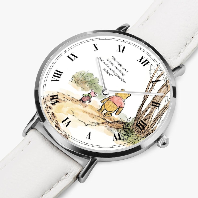 Pode incluir: Um rel&oacute;gio de pulso prateado com uma pulseira de couro branca. O mostrador do rel&oacute;gio apresenta uma ilustra&ccedil;&atilde;o em aquarela de Winnie the Pooh e Piglet, com algarismos romanos e a cita&ccedil;&atilde;o: "How lucky am I to have something that makes saying good-bye so hard."