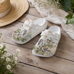 May include: White clogs with a nature-themed design of birds, berries, and foliage. The clogs have a strap for a secure fit. The design is printed on the front of the clogs. The clogs are on a wooden surface.