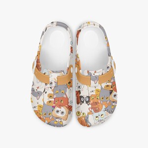 May include: A pair of white clogs covered in a colorful pattern of cartoon cats. The cats are in various poses and colors, including orange, gray, and brown. The clogs have tan straps and white interiors.
