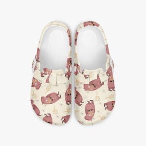 May include: A pair of cream-colored clogs with a repeating pattern of pink animals, daisies, and wheat stalks. The clogs have white straps and are designed for casual wear. The shoes are a size 7.