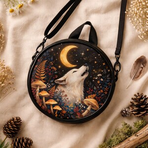 May include: Circular black crossbody bag with a wolf howling at a crescent moon against a starry night sky. The design includes mushrooms and foliage. It has a black strap and handle.