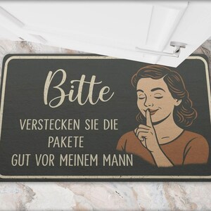May include: Black rectangular doormat with the German text "Bitte verstecken Sie die Pakete gut vor meinem Mann" in white script. A vintage-style illustration of a woman with a finger to her lips is on the right.