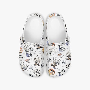 Puede incluir: Un par de zuecos blancos con un estampado de perro dálmata. El estampado presenta perros manchados en blanco y negro en varias poses, junto con estrellas y calabazas. Los zuecos tienen correas y suelas blancas.