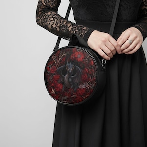 May include: Black round crossbody bag with a dragon illustration set against red roses. The bag has a black strap and body. The dragon is black with blue details. The background is a dark red floral pattern.