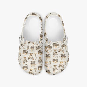 May include: A pair of white clogs with a repeating pattern of owls and wooden crates. The owls are in various poses, and the crates appear to hold eggs. The clogs have a rounded toe and a strap that can be worn around the heel.