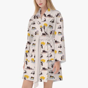 May include: A white robe with a repeating pattern of black cats and books in yellow, black, and white. The robe has a matching belt and a shawl collar. The robe is knee-length.
