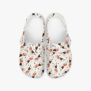 May include: A pair of white clogs with a repeating pattern of brown, black, and white Basset Hound illustrations and paw prints. The clogs have a strap that can be worn around the heel or over the top of the foot.