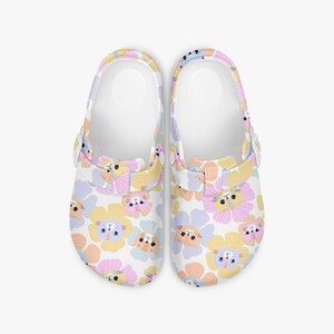 Pode incluir: Um par de tamancos brancos com um padrão floral colorido com rostos de gato. As flores são em tons de amarelo, rosa, laranja e azul. Os tamancos têm uma tira branca e um fecho de fivela.