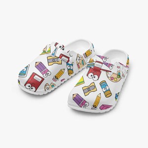 Puede incluir: Zuecos blancos con un colorido estampado de lápices de dibujos animados, cuadernos, sacapuntas y paletas de pintura. Los zapatos tienen un diseño de punta cerrada y una correa en la parte trasera para un ajuste seguro. Ideales para amantes del arte.