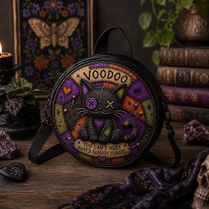 May include: A round black bag featuring a graphic of a stitched black cat with a purple button eye and an X-shaped eye. The design includes purple and orange patchwork patterns, the text VOODOO, and the phrase YOU CAN'T HURT WHAT'S ALREADY CURSED.