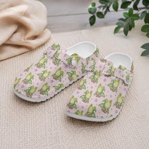 May include: A pair of pink clogs with a repeating pattern of green cartoon alligators and purple stars. The clogs have white soles and adjustable straps.