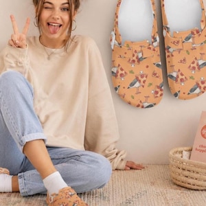 May include: Orange clogs with a pizza and bird pattern. The clogs are shown with a person wearing a beige sweatshirt and blue jeans. The clogs are on a rug.