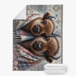 Könnte beinhalten: Eine dekorative Decke mit einer Illustration von zwei Bisonkälbern, die in gemusterte Decken gehüllt sind. Die Bisons haben braunes Fell und dunkle Hörner, wobei die Decken eine Mischung aus Farben und geometrischen Designs aufweisen. Eine gefaltete weiße Decke ist ebenfalls sichtbar.