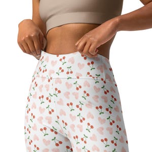 May include: White leggings with a repeating pattern of red cherries and pink hearts. The leggings have a high waistband and are made of a stretchy material. The overall theme is playful and feminine.