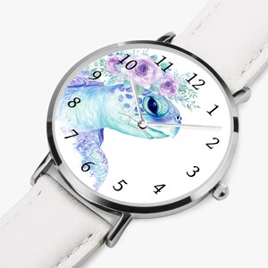 Könnte beinhalten: Weiße Armbanduhr mit silbernem Gehäuse und weißem Lederarmband. Das Zifferblatt zeigt eine Aquarellillustration einer Meeresschildkröte mit einem Blumenschmuck in Blau, Lila und Grün.