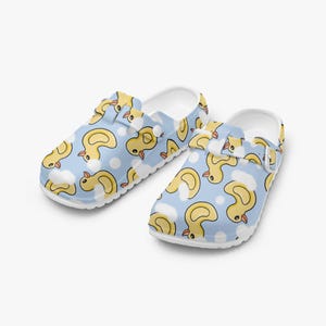 May include: A pair of white clogs with a light blue background and a pattern of yellow ducklings and white clouds. The clogs have adjustable straps and a slip-on design.