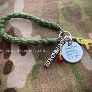 Può includere: Braccialetto in paracord verde con ciondoli argentati, tra cui un ciondolo con il testo "One Day Closer... One Day Stronger" e un ciondolo con il testo "Air Force". Il braccialetto presenta anche un ciondolo a forma di nastro giallo.
