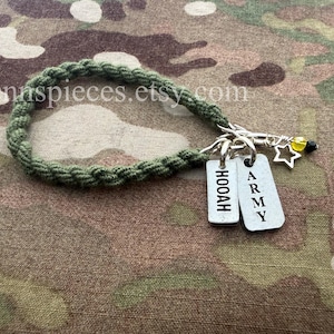 Puede incluir: Un llavero trenzado verde con dos etiquetas plateadas que dicen "HOOAH" y "ARMY". El llavero también presenta un dije de estrella y una cuenta amarilla y negra. El fondo es de tela de camuflaje.
