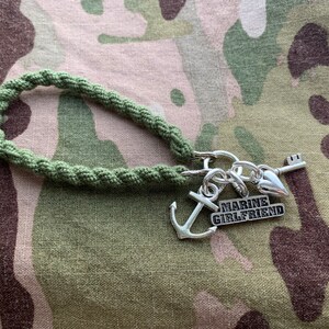 May include: A green braided cord keychain with a silver anchor charm, a silver heart charm, a silver key charm, and a silver charm with the text "MARINE GIRLFRIEND".